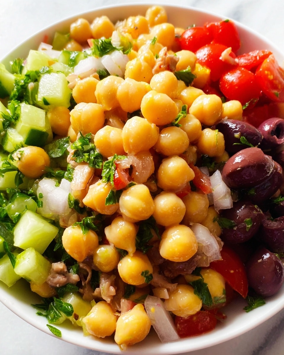 A clear glass container holds a colorful chickpea salad with many mix-ins. The main layer is golden-yellow chickpeas, large and round, filling most of the container. Mixed evenly throughout are chopped pieces of green cucumber, small purple-red onion cubes, bright red tomato chunks, and dark black olives. Flakes of light brown tuna are scattered between, and small green parsley leaves add freshness. A silver spoon sticks into the salad from the top left corner. The whole container sits on a white marbled surface. photo taken with an iphone --ar 4:5 --v 7