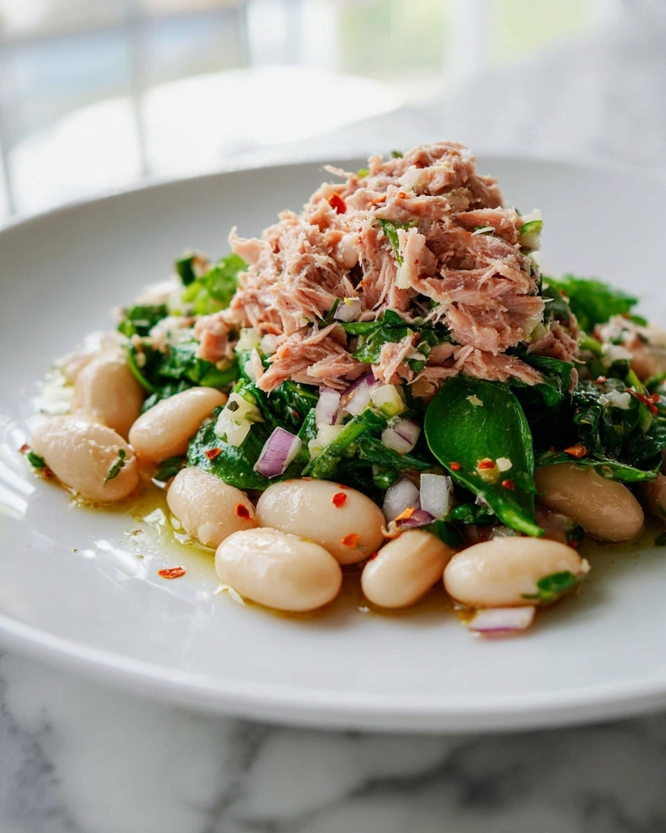 A white plate holds a fresh salad with two main layers. The bottom layer is made of large white beans mixed with bright green leafy herbs and small pieces of chopped purple onion, all glistening lightly with olive oil and seasoning. The top layer is a loose heap of shredded pinkish tuna, also mixed with some of the green herbs and finely chopped onion bits, creating a textured and colorful contrast. The beans look soft and smooth, while the tuna is flaky and moist, with a few red pepper flakes sprinkled on top. The whole dish sits on a white marbled surface, with soft light from the background. photo taken with an iphone --ar 4:5 --v 7