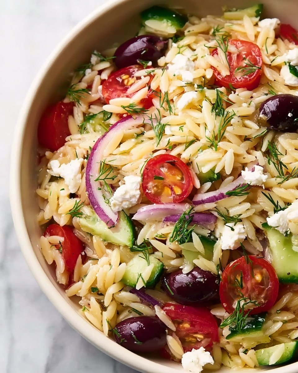 A close-up view of a white bowl filled with a colorful orzo salad, showing a mix of small, pale yellow orzo pasta grains as the base layer. Scattered throughout are sliced bright red cherry tomatoes, deep purple kalamata olives, chunks of green cucumber, and thin slices of purple-red onion, creating varied pops of color. Small white crumbles of feta cheese are spread evenly on top, along with sprigs of fresh green dill adding a light texture contrast. The bowl is placed on a white marbled surface. photo taken with an iphone --ar 4:5 --v 7